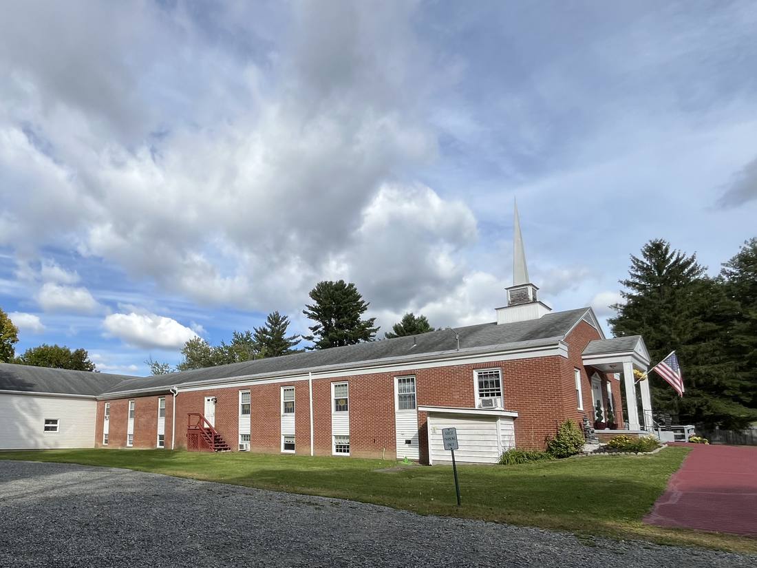 The long red brick side of the church and its white steeple, seen across the gravel parking lot and the lawn under a wide sky with clouds. The columned front entry and a flag are at the right.