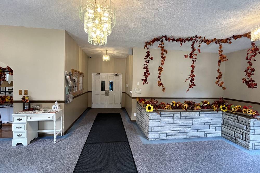 The entry lobby: a hallway to a pair of white doors with a dark mat on the carpet, a glass chandelier, and a stone-faced planter filled with autumn flowers.